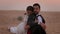 Portrait Of Young Couple In The Desert. They Are Sitting On The Sand. Woman Huddles Behind The Shoulder Of Man.