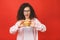 Portrait of young curly beautiful hungry woman eating burger. Isolated portrait of student with fast food over red background.