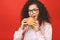 Portrait of young curly beautiful hungry woman eating burger. Isolated portrait of student with fast food over red background.