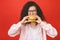 Portrait of young curly beautiful hungry woman eating burger. Isolated portrait of student with fast food over red background.