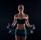 Portrait of a young fitness woman in sportswear doing workout with dumbbells on black background. Tanned athletic girl.