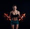 Portrait of a young fitness woman in sportswear doing workout with fiery dumbbells on black background. Tanned athletic girl.