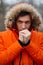 Portrait of young frozen man in hood and orange jacket. Close up of adult male warming hands with his breath in cold