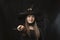 Portrait of young girl in witchs costume wears pointed hat with magic wand in her hands on black background