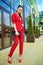 Portrait of young gorgeous lady with high pony tail in red costume and high-heeled shoes standing in front of mirrored shop window