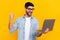 Portrait of young handsome guy holding laptop fist up celebrates good marks online education isolated on yellow color