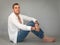 Portrait of a young handsome man dressed in a white unbuttoned shirt and blue jeans sitting on floor in studio on grey