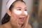 Portrait of young happy and beautiful Asian Chinese woman applying facial anti panda eyes patch at bathroom mirror in skin care