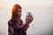 Portrait of young happy woman in hipster shirt standing in front of picturesque sea view, taking selfie with sunset or