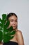 Portrait of a young magnificent half-naked asian girl in a black top hiding under a monstera leaf