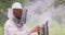 Portrait young, male bearded beekeeper in white protective suit closes a lid of smoking smoker, which is on the hive