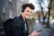 Portrait of young man commuter with headphones standing outdoors in city, using smartphone.