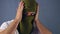 Portrait of a young man putting on a military fabric mask over his head against a gray background. Medium shot.