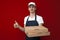 Portrait of a young pizza delivery man in uniform on a red background, a courier guy gives a box of pizza and shows like