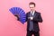 Portrait of young positive magician, man in elegant suit standing pointing at big blue fan. indoor studio shot isolated on pink