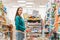 Portrait of a young pretty Caucasian woman in casual clothing poses with a grocery cart in the aisle of a store. Copy space. The