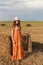 Portrait of a young rural woman in a sarafan and hat that stands near a haystack in a wheat field with a stormy sky