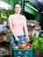 Portrait of young satisfied female purchaser holding shopping basket with fruits and vegetables