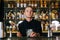 Portrait of young serious bartender standing behind bar counter and holding stirring spoon and glass filled with ice