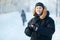 Portrait of a young Siberian man with phone in hands, wearing warm down jacket, fur hood. Cold winter day