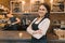 Portrait of young smiling female cafe worker, standing at the counter. Woman with folded hands, professional baristas team cafe