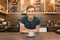 Portrait of young smiling male barista with prepared drink with arms crossed standing behind cafe counter. Coffee shop business co