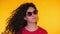 Portrait of young spanish girl with curls puts on sunglasses on yellow background. Tempting woman smiling to camera