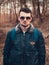 Portrait of young trendy man in blue aviator bomber jacket with sunglasses  standing on the path in forest.  Fashion male model