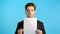 Portrait of young unhappy serious businessman in suit holding white vertical a4 paper isolated on blue studio background