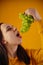 Portrait of young woman with bunch of grapes on yellow background. Close up of female opens mouth, going to eat green