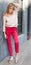 Portrait of a young woman dressed in a blouse, red Chino Trouser, and gold brogues. Posing next to a gray wall. Fashion.