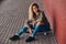 Portrait of a young woman dressed in a hoodie and ripped jeans sitting on a skateboard at bridge footway.