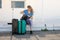 Portrait of young woman with fair hair sitting on white wooden floor of veranda rummaging in black bag. Travelling.