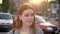 Portrait of young woman with ginger hair standing in street and looking in camera during evening, smiling, traffic of