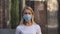 Portrait of young woman in medical mask looking at camera. Blonde female in a white t-shirt standing outdoors on blurred