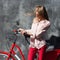 Portrait young woman in red chino holding hands on her red vintage bicycle, standing against gray wall. The wind blows her hair. O