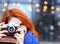 Portrait of young woman with red hair takes pictures of landmarks in the city in a short coat and a knitted blue yoke. Sunbeams