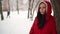 Portrait of young woman in red jacket and hood in winter season. Beautiful lady stands near tree and looks away. Fluffy