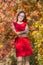 Portrait of young woman in red short dress in fall forest. Female person with bouquet from autumn leafs posing embracing himself