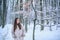 Portrait of a young woman in snow trying to warm herself. Beautiful girl in the winter forest in white down jacket.