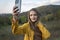 Portrait of Young woman taking selfie on smartphone, mountain background. Girl in yellow jacket enjoys of autumn nature