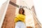 Portrait of a young woman with wavy hair, wear in yellow sweater and white jeans behind modern building with cross arm