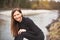 Portrait of young woman wearing black jeans and top, leaning, her long hair falling down, smiling. Blurred river in background