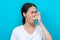 Portrait of a young woman wincing when using an inhaler. Blue background. Copy space