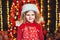 Portraite of little curly blonde girl in Santa hat on blurred lights background. Christmas concept