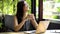 Positive Asian female student in glasses smiling and browsing laptop while sitting at table on terrace and studying on