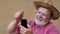 Positive bearded senior man in straw hat listening music from cellular phone