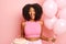 Positive brunette young woman poses with balloons and birthday cake with burning candles,  over pink background