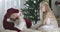 Positive Caucasian girl with blond curly hair making hush gesture and talking to Santa at home. Child spending Christmas