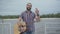 Positive Caucasian man standing with guitar on embankment and waving. Portrait of happy young handsome musician looking